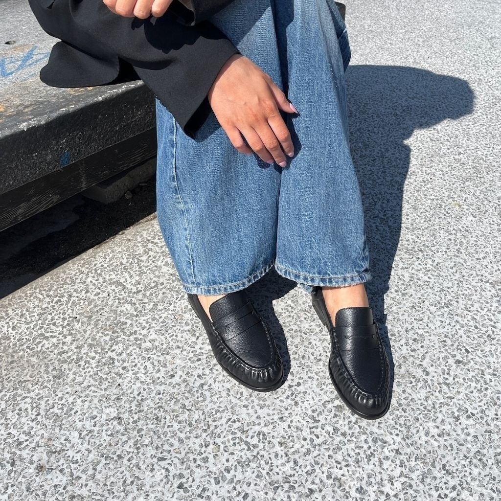 ShoeShop black leather loafers with a classic penny strap design worn with blue jeans on a textured outdoor surface