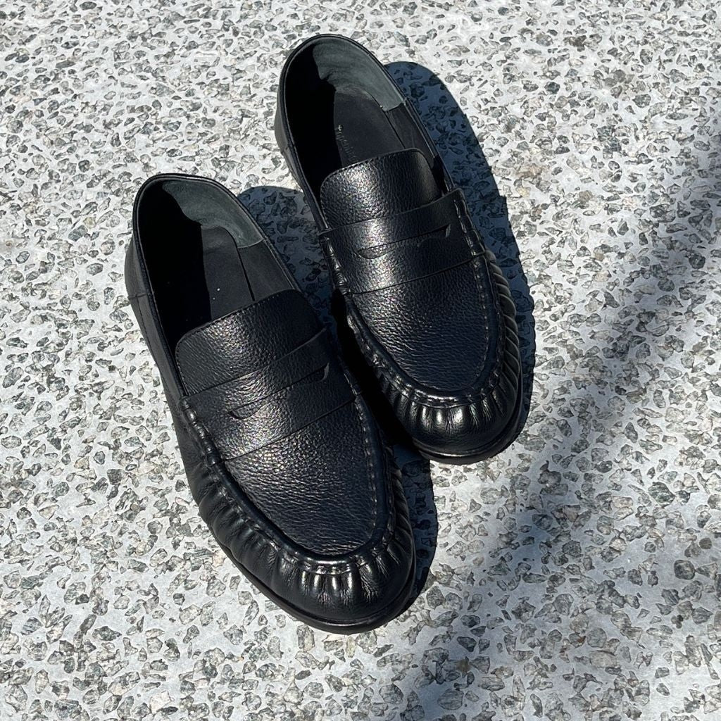 ShoeShop black leather penny loafers with a textured finish and moccasin-style stitching on a stone surface