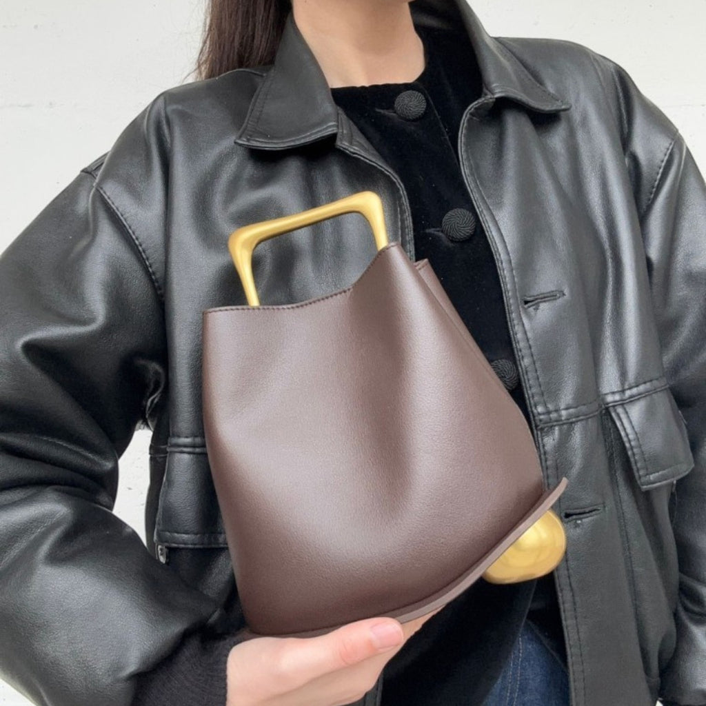 ShoeShop brown structured handbag with a unique gold metal handle held by a person wearing a black leather jacket