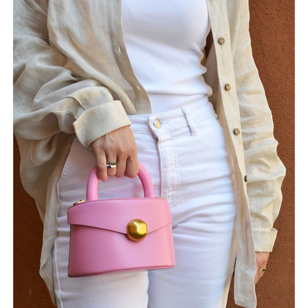 ShoeShop pink leather handbag with a gold magnetic button closure, held by a woman in white jeans and linen shirt.